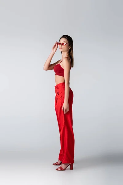 Full length of young woman in red outfit adjusting sunglasses on grey — Stock Photo