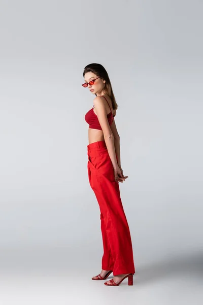 Full length of brunette woman in sunglasses and red outfit posing on grey — Stock Photo