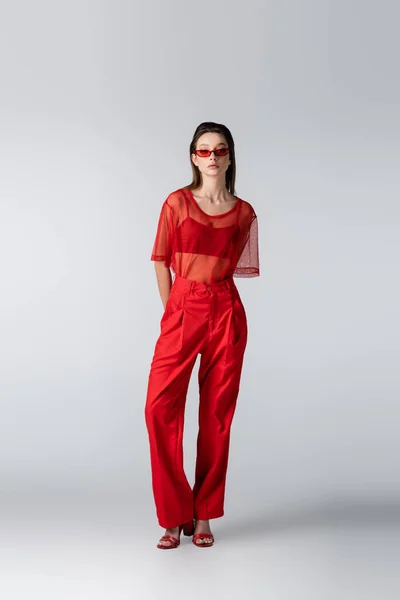 Tutta la lunghezza del modello giovane in occhiali da sole e abito rosso alla moda in posa sul grigio — Foto stock