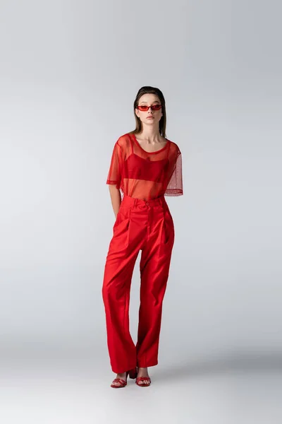 Longueur totale de jeune modèle en lunettes de soleil et tenue tendance rouge posant sur le gris — Photo de stock