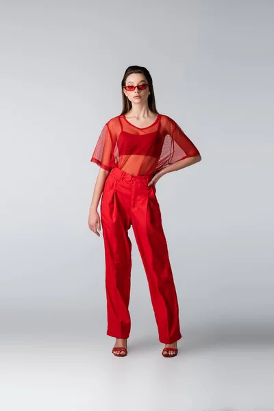 Longitud completa de modelo joven en gafas de sol y traje rojo de moda posando con la mano en la cadera en gris - foto de stock