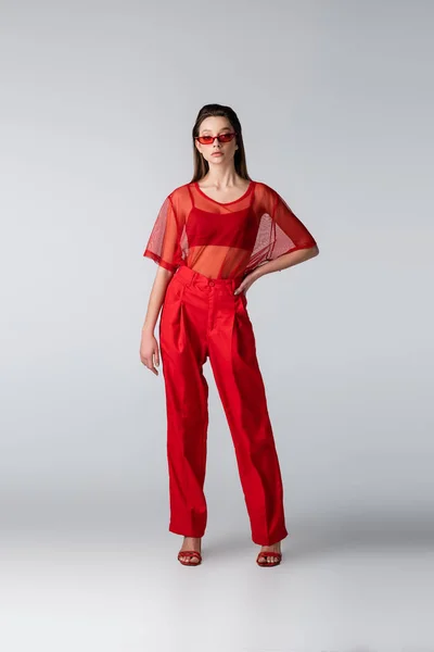 Toute la longueur du jeune modèle en lunettes de soleil et tenue tendance rouge posant avec la main sur la hanche sur gris — Photo de stock