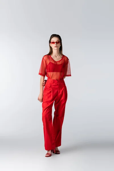 Longueur totale de jeune modèle en tenue rouge tendance et lunettes de soleil posant sur le gris — Photo de stock