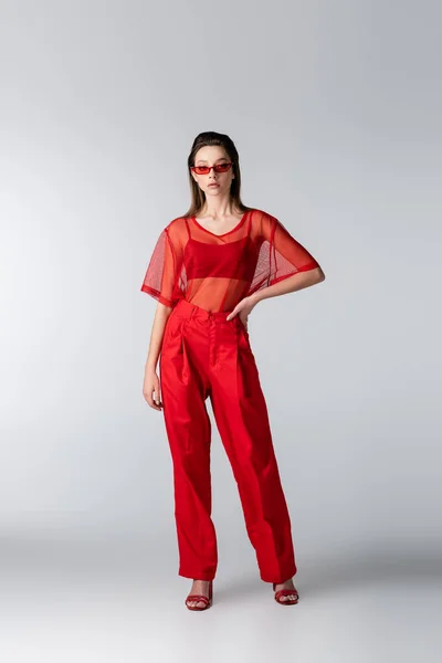 Longueur totale de jeune modèle en tenue rouge tendance et lunettes de soleil posant à la main sur la hanche sur gris — Photo de stock