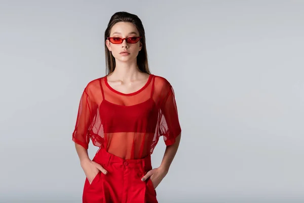 Молода модель в червоному модному вбранні і сонцезахисних окулярах позує руками в кишенях ізольовані на сірому — стокове фото
