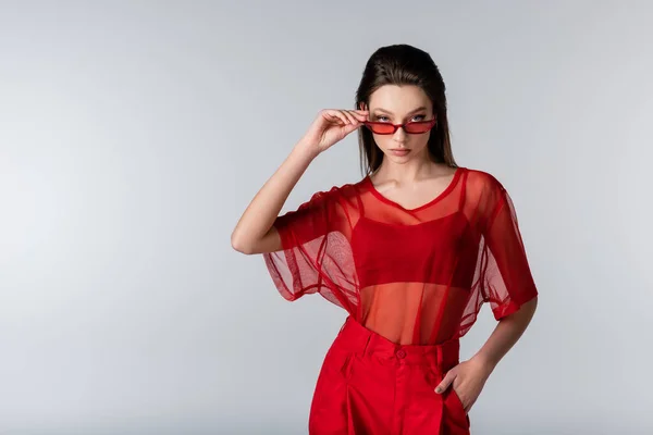 Молода модель в червоному модному вбранні, що регулює сонцезахисні окуляри, позує руками в кишені ізольовано на сірому — стокове фото
