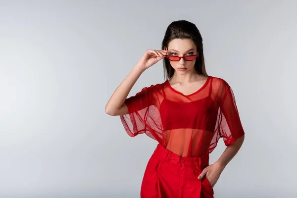 Modelo joven en traje de moda rojo ajustando gafas de sol mientras posa con la mano en el bolsillo aislado en gris - foto de stock