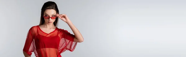 Young model in red trendy outfit adjusting sunglasses while posing isolated on grey, banner — Stock Photo