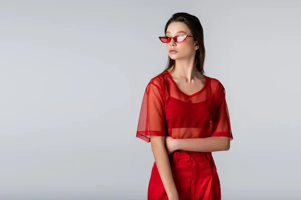 Jeune modèle en tenue tendance et lunettes de soleil rouges posant isolées sur gris — Photo de stock