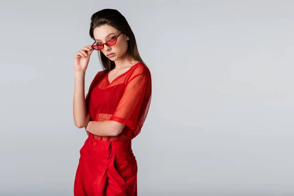 Jeune femme en tenue rouge tendance ajustant les lunettes de soleil tout en posant isolé sur gris — Photo de stock