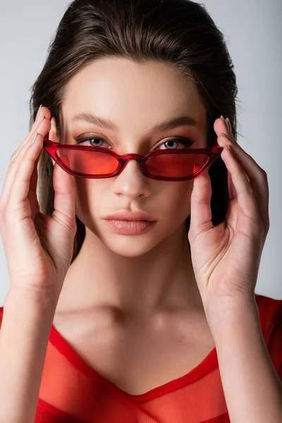 Modelo joven ajustando gafas de sol rojas de moda aisladas en gris - foto de stock