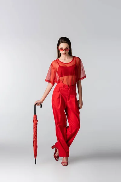 Longitud completa de modelo joven en traje de moda rojo y gafas de sol posando con paraguas en gris - foto de stock