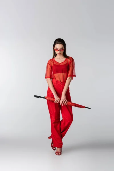 Longueur totale de jeune modèle en tenue rouge tendance et lunettes de soleil tenant parapluie sur gris — Photo de stock