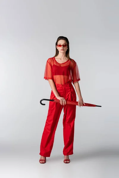 Longitud completa de la mujer joven en traje de moda rojo y gafas de sol con paraguas en gris - foto de stock