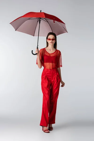Полная длина молодой женщины в красном модном наряде и солнцезащитных очках, стоящих под зонтиком на сером — стоковое фото