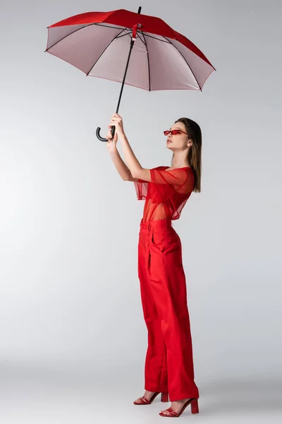Comprimento total do modelo jovem em roupas vermelhas da moda e óculos de sol segurando guarda-chuva acima da cabeça em cinza — Fotografia de Stock