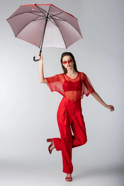 Piena lunghezza del modello giovane in abito alla moda e occhiali da sole rossi in posa sotto l'ombrello su grigio — Foto stock