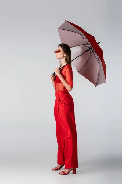 Comprimento total da jovem mulher na roupa da moda e óculos de sol de pé com guarda-chuva vermelho no cinza — Fotografia de Stock