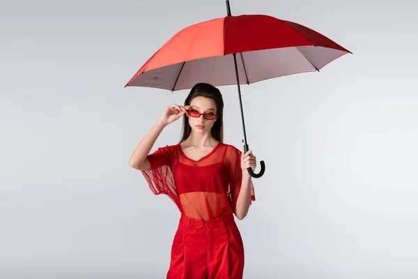 Young woman in trendy outfit adjusting red sunglasses and standing under umbrella isolated on grey — Stock Photo