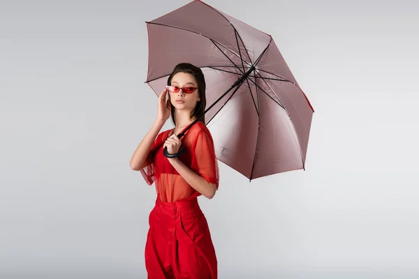 Junge Frau im roten Outfit mit Sonnenbrille und Schirm isoliert auf grau — Stockfoto