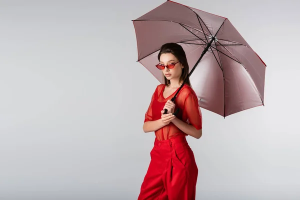 Young woman in red outfit and sunglasses standing under umbrella isolated on grey — Stock Photo