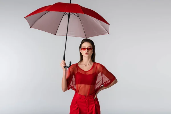 Jeune femme en tenue tendance et lunettes de soleil rouges debout avec la main sur la hanche sous parapluie isolé sur gris — Photo de stock