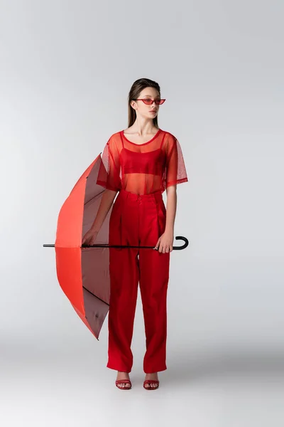 Pleine longueur de jeune femme en tenue rouge tendance et lunettes de soleil tenant parapluie sur gris — Photo de stock