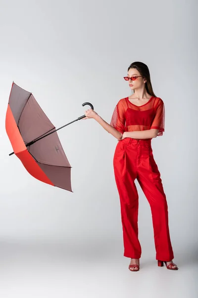 Piena lunghezza di giovane donna in abito rosso alla moda e occhiali da sole in piedi con ombrello su grigio — Foto stock