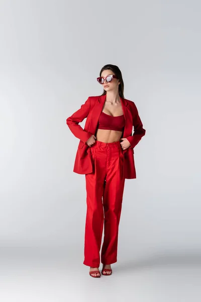 Повна довжина молодої жінки в модному червоному костюмі з верхньою частиною культури і сонцезахисними окулярами, що стоять на сірому — стокове фото