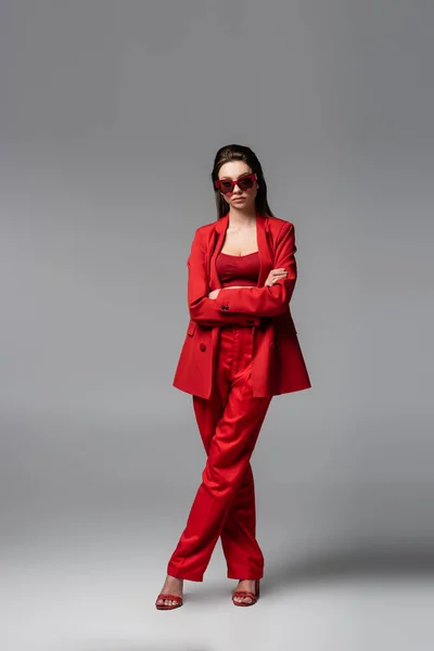 Piena lunghezza di giovane donna in abito rosso alla moda e occhiali da sole in piedi con braccia incrociate su grigio scuro — Foto stock