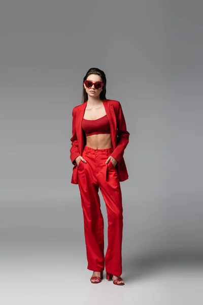 Longueur pleine de jeune femme en costume rouge tendance et lunettes de soleil debout avec les mains dans les poches sur gris foncé — Photo de stock