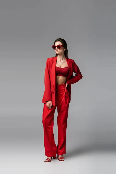 Comprimento total da jovem mulher em terno vermelho na moda e óculos de sol de pé com a mão no quadril no cinza escuro — Fotografia de Stock