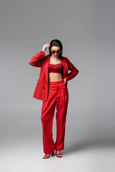 Повна довжина молодої моделі в червоному костюмі і сонцезахисних окулярах, що стоять з рукою на стегні на темно-сірому — стокове фото