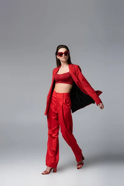 Piena lunghezza di giovane donna in abito rosso alla moda e occhiali da sole che camminano su grigio scuro — Foto stock