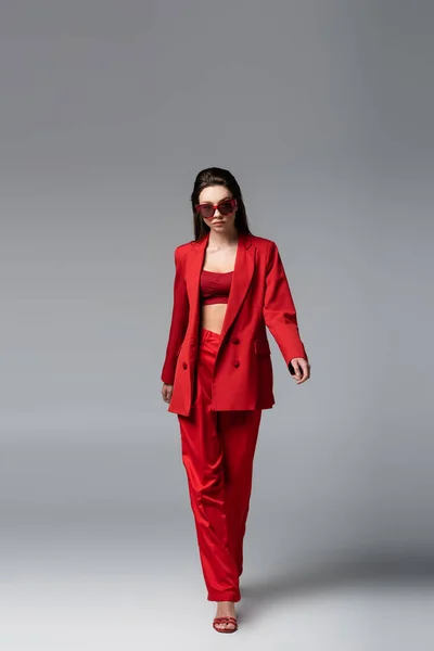 Full length of young woman in red suit and trendy sunglasses walking on dark grey — Stock Photo