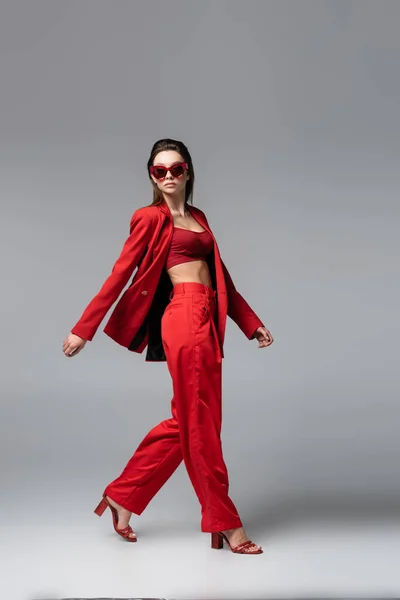 Full length of brunette woman in red suit and trendy sunglasses walking on dark grey — Stock Photo