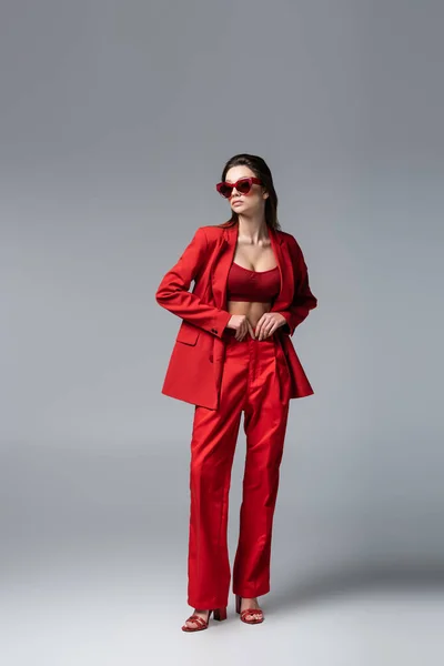 Piena lunghezza di giovane donna in abito rosso alla moda e occhiali da sole in posa su grigio scuro — Foto stock