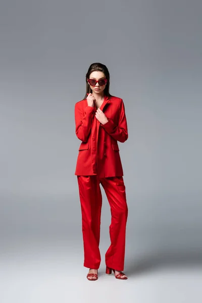 Comprimento total da jovem mulher em terno vermelho e óculos de sol em pé no cinza escuro — Fotografia de Stock