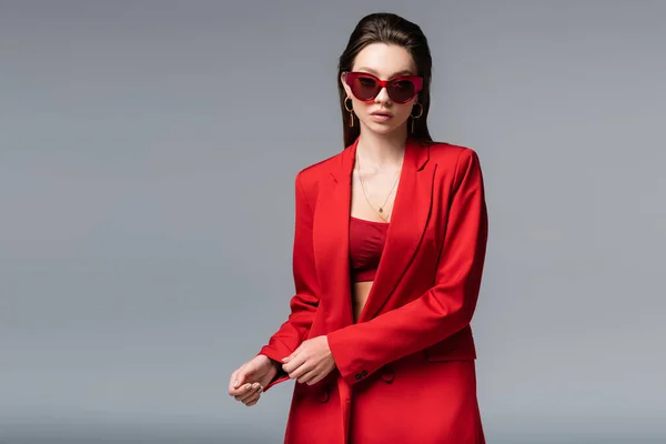 Jeune femme en costume rouge tendance et lunettes de soleil posant isolées sur gris foncé — Photo de stock