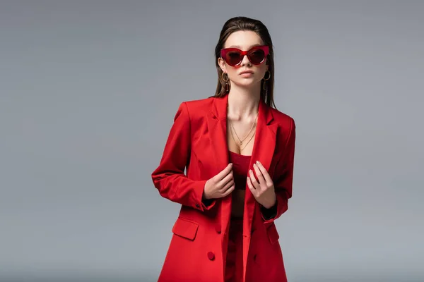 Молода модель в модному червоному костюмі і сонцезахисних окулярах позує ізольовано на темно-сірому — стокове фото