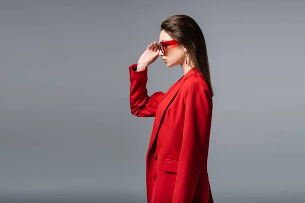 Vista lateral de mujer joven en traje rojo de moda ajustando gafas de sol aisladas en gris oscuro - foto de stock