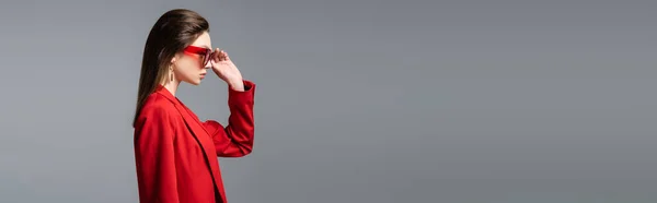 Vista laterale della giovane donna in abito rosso alla moda che regola gli occhiali da sole isolati su grigio scuro, banner — Foto stock