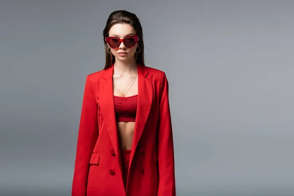 Молода жінка в модному червоному костюмі з верхнім і сонцезахисними окулярами дивиться на камеру ізольовано на темно-сірому — стокове фото