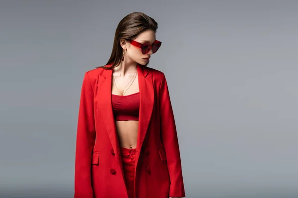 Молода жінка в червоному костюмі з верхнім і сонцезахисними окулярами дивиться ізольовано на темно-сірому — стокове фото