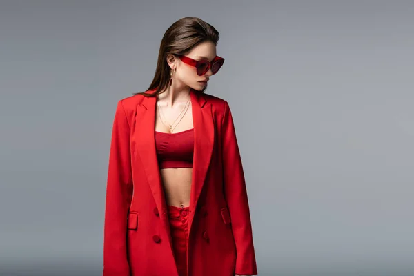 Jeune femme en costume rouge avec haut de culture et lunettes de soleil regardant loin isolé sur gris foncé — Photo de stock