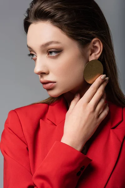 Trendy giovane donna in giacca rossa toccante orecchino isolato su grigio — Foto stock