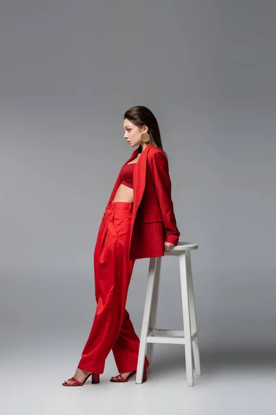 Junge Frau im trendigen roten Anzug steht neben weißem Stuhl auf dunkelgrau — Stockfoto