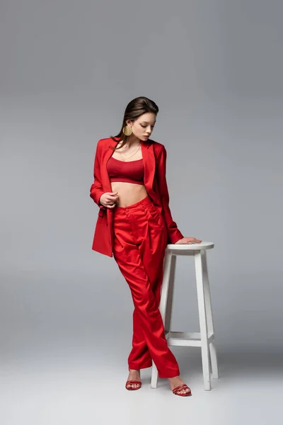 Longitud completa del modelo joven en traje rojo de moda de pie cerca de la silla blanca en gris oscuro - foto de stock