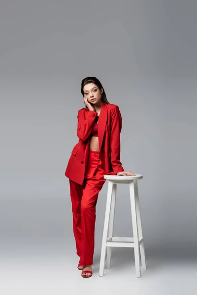 Piena lunghezza di giovane donna in abito rosso alla moda in posa vicino sedia bianca su grigio scuro — Foto stock