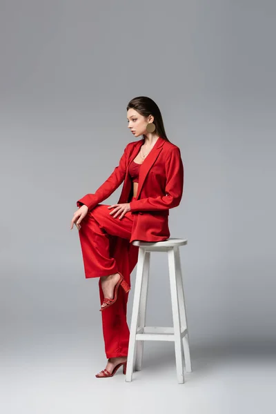 Pleine longueur de jeune femme en costume rouge tendance assis sur une chaise blanche sur gris foncé — Photo de stock